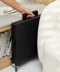 Compact black folding utility cart with lid and basket on four 360 degree rotating wheels, stored upright under a table.