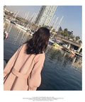 Woman wearing a pink coat stands by the water, overlooking boats at a marina with urban buildings in the background.
