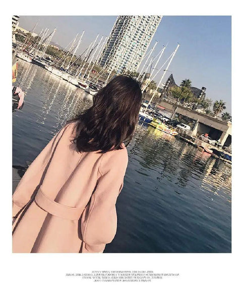 Woman wearing a pink coat stands by the water, overlooking boats at a marina with urban buildings in the background.