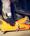 Orange hydraulic floor jack lifting a car; gloved hand near wheel and pinch-weld pad