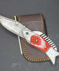 Damascus Steel Folding Knife With Ladder Pattern Blade And Copper Fish Style Handle resting on leather sheath.