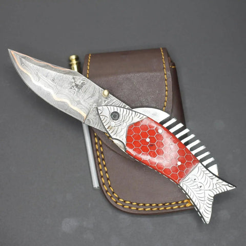 Damascus Steel Folding Knife With Ladder Pattern Blade And Copper Fish Style Handle resting on leather sheath.
