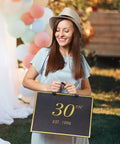 Woman holding 30th birthday black and gold gift bag with '30th Est. 1996' text, outdoors with balloons in background.