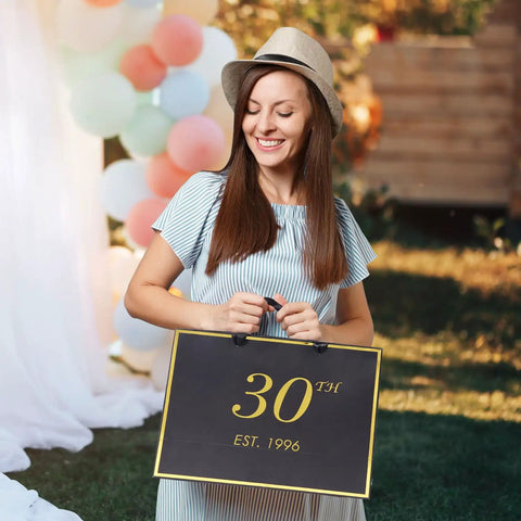 Woman holding 30th birthday black and gold gift bag with '30th Est. 1996' text, outdoors with balloons in background.