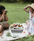Women enjoying a picnic with a gray Collapsible Wash Basin With Drain Plug 2.4 Gallon Foldable Kitchen Sink filled with drinks.