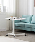 Slick Mobile Lectern Podium Stand With Smooth Roll Sit Stand Laptop Desk And Roomy Notebook Cart Build For Home Office Flex Workstations In Clean White Finish next to a couch.