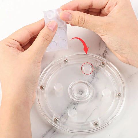 Hands applying rubber feet to the clear acrylic turntable from the Level Up Your Sweet Baking Game With This Solid Acrylic Cookie Decorating Turntable Kit That Comes Loaded With Scribe Needles Soft Brushes Grip Mats And Rubber Feet For Flawless Icing Every Time.