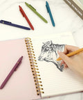Hand drawing in notebook using 6 Pack 0.5mm Gel Pens Quick Dry Black Ink, Fine Point Rollerball Pens, office desk essentials.