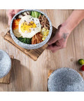 Person serving bibimbap in Granite Stone Bowl With Wooden Base For Bibimbap And Korean Stews, 24oz size, on wooden table.