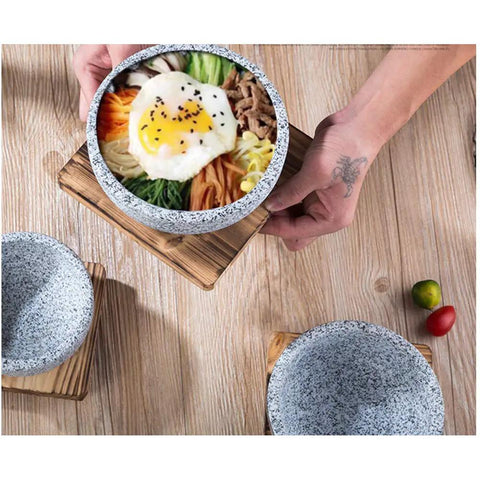 Person serving bibimbap in Granite Stone Bowl With Wooden Base For Bibimbap And Korean Stews, 24oz size, on wooden table.
