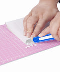 Blue retractable utility knife cutting white paper on pink cutting mat.