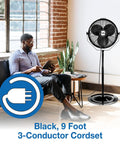 18 Inch Industrial Pedestal Fan With Powder Coated Steel Frame Delivers Beastly Airflow For Workshops And Garages Featuring A One Sixth Horsepower Enclosed Motor And A Ten Foot SJT Cord Plus A One Year Limited Warranty near a seated man in a room.