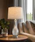 24.5 Inch Glass Table Lamps Set Of 2 With USB Charging Ports, Nightstand Bedside Lamps For Living Room Bedroom Office Or College, Modern Silver Finish, Built In 5V/2A USB Ports That Charge Your Phone On Or Off on a wooden table by a sofa.
