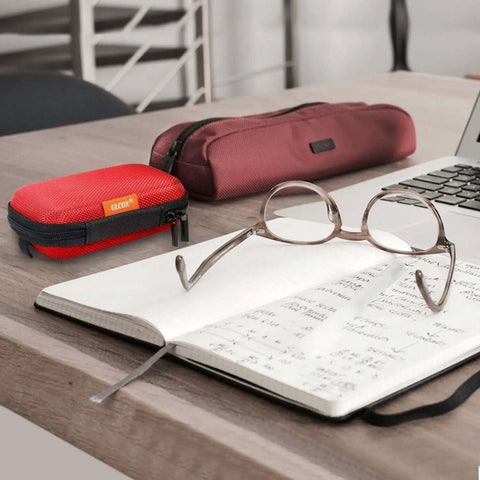 Small Red Rectangular Eva Case With Hella Tough Crush Proof Hard Shell And Smooth Zipper Mesh Pouch For Charger Cables Earbuds Memory Cards And Everyday Carry on a desk beside glasses and a notebook.
