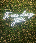 Neon sign on greenery wall reading 'It was always you.'