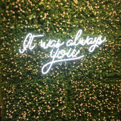 Neon sign on greenery wall reading 'It was always you.'