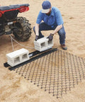 Heavy Duty Steel Drag Harrow And Field Leveler For ATV UTV And Tractor With Front Tray 5 By 4 Feet Keeps Soil Even For Lawn Farm And Landscape Prep For Soil Leveling And Dirt Moving On Fields Or Driveways, Easy To Attach And Use setup by a person hooking it to an ATV on a sandy field.