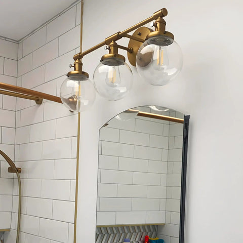 Three Light Vintage Industrial Bathroom Vanity Wall Sconce With Handblown Clear Glass Globes And Antique Gold Finish, 25.2 Inch Wide Triple Light Bath Fixture For A Soft Industrial Look shown above a mirror in a bathroom