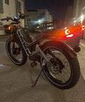 Romeo Pro Electric Bike With 5600W Dual Motors Hitting Fifty Mph Shredding Gnarly Dirt Paths Using Sixty Volt Eighty Amp Hours Cells Yielding Two Hundred Fifty Miles Across Chunky Four Point Zero Foot Tires And Rigid Rear Cargo Bags in urban setting at night.