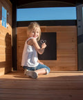 Child drawing on chalkboard inside Cedar Wood Swing Set With Wave Slide And Elevated Clubhouse Play Kitchen And Picnic Area