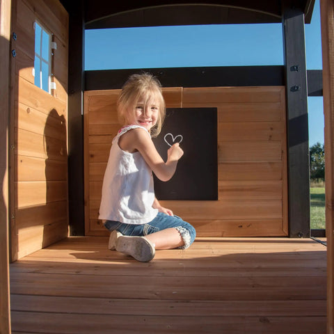 Child drawing on chalkboard inside Cedar Wood Swing Set With Wave Slide And Elevated Clubhouse Play Kitchen And Picnic Area