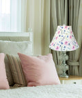 Floral lamp shade on glass table lamp beside bed with pink pillows, in cozy bedroom setting with curtains.