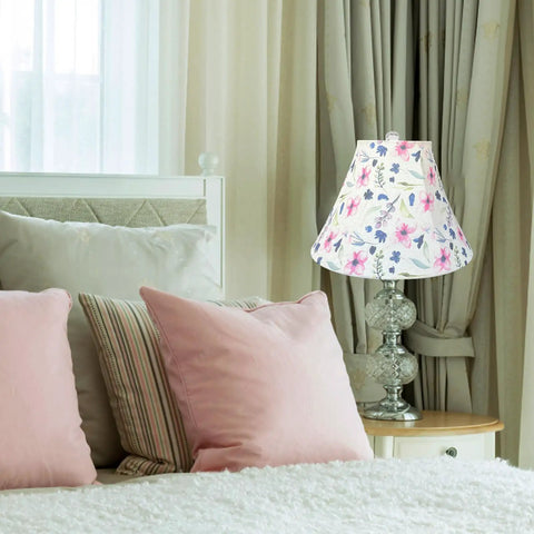Floral lamp shade on glass table lamp beside bed with pink pillows, in cozy bedroom setting with curtains.