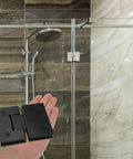 Matte black 304 stainless steel shower door hinge held in hand, bathroom background, for frameless doors, 180-degree clamp