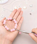 Person using pliers to attach a pink beaded bracelet with silver charms, surrounded by silver jewelry components on a table.