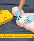 AED trainer device (yellow) with CPR training mannequin and electrode pad being applied by blue-gloved hands.