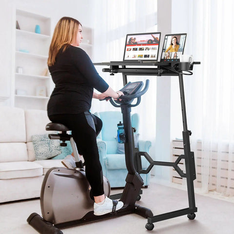 Woman using exercise bike next to small standing desk on wheels for treadmill workouts, portable rolling desk holding two tablets.