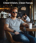 Front view of man in cafe wearing sunglasses, seated at a wooden table with laptop and coffee cup.