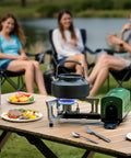 Portable Butane Camping Stove With Foldable Bucket Design Compact Gas Burner For Camping Emergency Use And Outdoor Cooking Sturdy Pot Support Carry Bag Included in use on a wooden outdoor table with people chatting in the background, showing cooking in action.