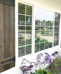 Exterior view of window with wooden shutters, showing purple flowers below and a suburban street in background.