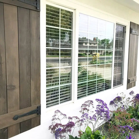 Exterior view of window with wooden shutters, showing purple flowers below and a suburban street in background.