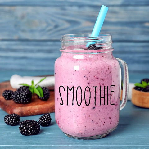 Glass jar with blackberry smoothie and straw, surrounded by fresh blackberries on wooden cutting board with the word 'SMOOTHIE' on the jar.