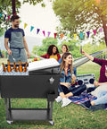 80 Quart Rolling Cooler Cart On Wheels With Dual Tops Built In Bottle Opener And Cap Catch Handy Shelf Portable Bar For Outdoor Patio Party And Backyard BBQ Ready In Black Keeps Ice Cold Up To 48 Hours With Ice Sheet in use at a picnic with people.