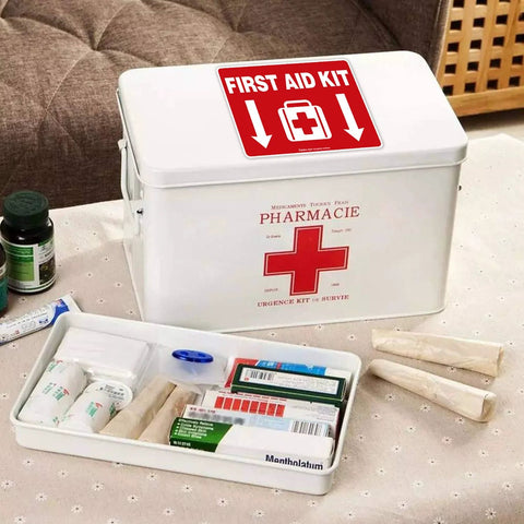 Front view of a white metal first aid kit with red FIRST AID KIT decal on lid; medical supplies nearby.