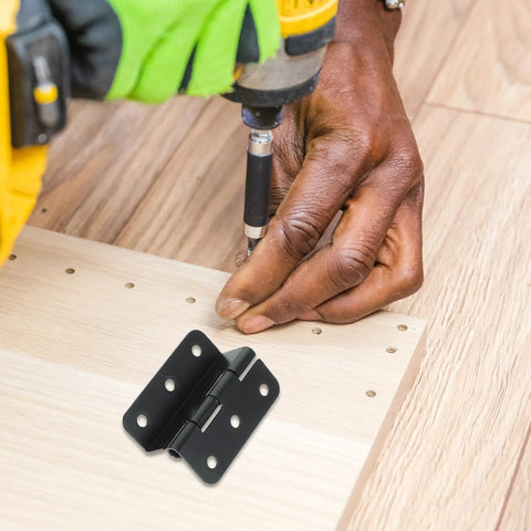 Close-up of a black iron cabinet box hinge being installed on light wood board with a drill nearby.