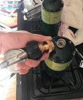 Hand holding a brass Propane Tank Adapter 20lb To 1lb Universal Converter For Disposable Cylinders next to camp stove