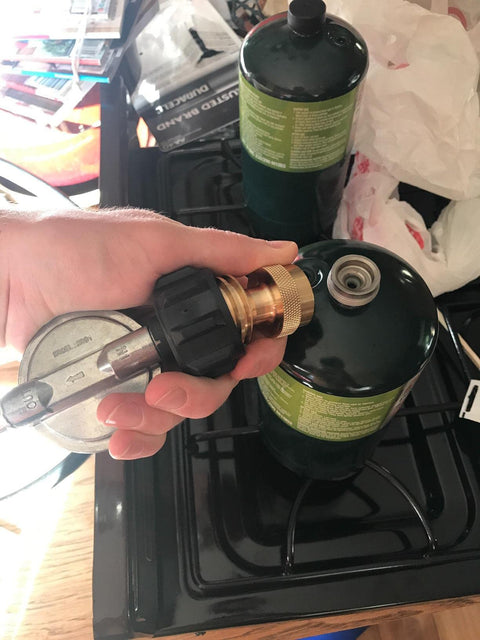 Hand holding a brass Propane Tank Adapter 20lb To 1lb Universal Converter For Disposable Cylinders next to camp stove