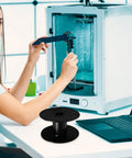 Super Slick Empty Cable Winder Reel 165mm Plastic Cord Organizer on table while a woman uses caliper near a 3D printer.