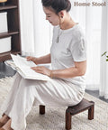 Woman sitting on a wooden home stool reading a book, wearing white attire, in a softly lit room.