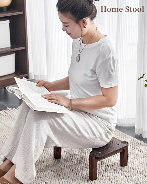 Woman sitting on a wooden home stool reading a book, wearing white attire, in a softly lit room.