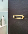 Brass bathroom door sign on dark wood, handcrafted metal decor
