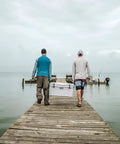 Two men walking on a dock with Heavy Duty 65 Quart Rotomolded Hard Shell Cooler Locks In Frost All Day Long To Keep Cold Brews And Fresh Catch Chilled For Tailgates Or Summer Getaways In Canopy Green.