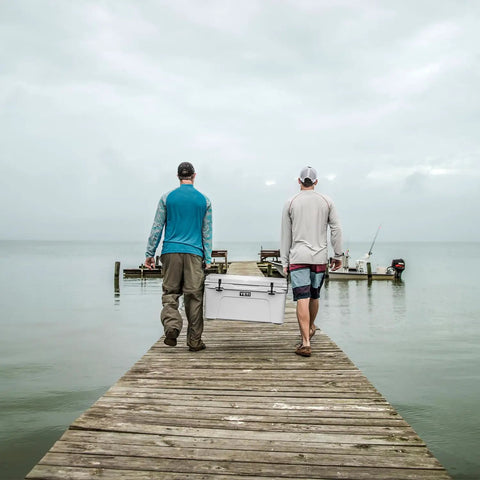 Two men walking on a dock with Heavy Duty 65 Quart Rotomolded Hard Shell Cooler Locks In Frost All Day Long To Keep Cold Brews And Fresh Catch Chilled For Tailgates Or Summer Getaways In Canopy Green.