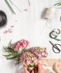 Hands arranging flowers using Must Have All In One Floral Tape And Wire Toolkit For DIY Bouquets And Arrangements on white surface.