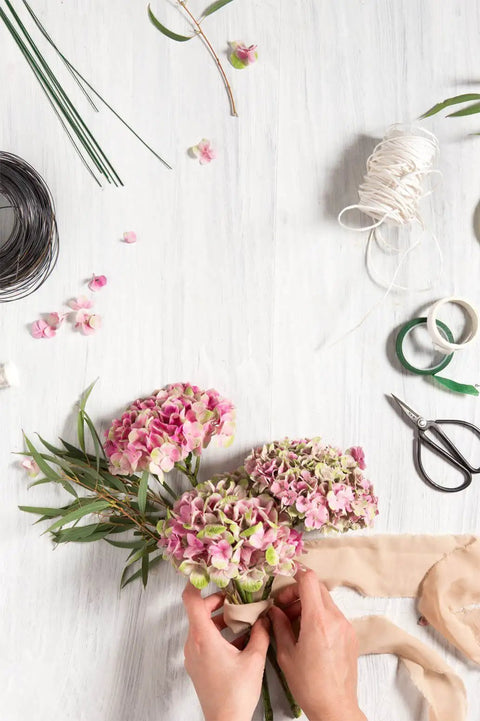 Hands arranging flowers using Must Have All In One Floral Tape And Wire Toolkit For DIY Bouquets And Arrangements on white surface.