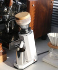 Kickstart Your Daily Grind With This Rock Solid Electric Coffee Grinder Featuring 40Mm Conical Titanium Burrs 19 Straight Forward Settings And A Generous 75G Bean Hopper. Image shows a white coffee grinder with wooden accents, a glass brewing setup, and a stainless steel cup.